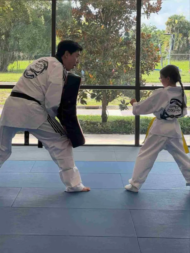 Young martial arts student practicing a punch while an instructor holds a training pad in class, learning self defense principles for beginners.
